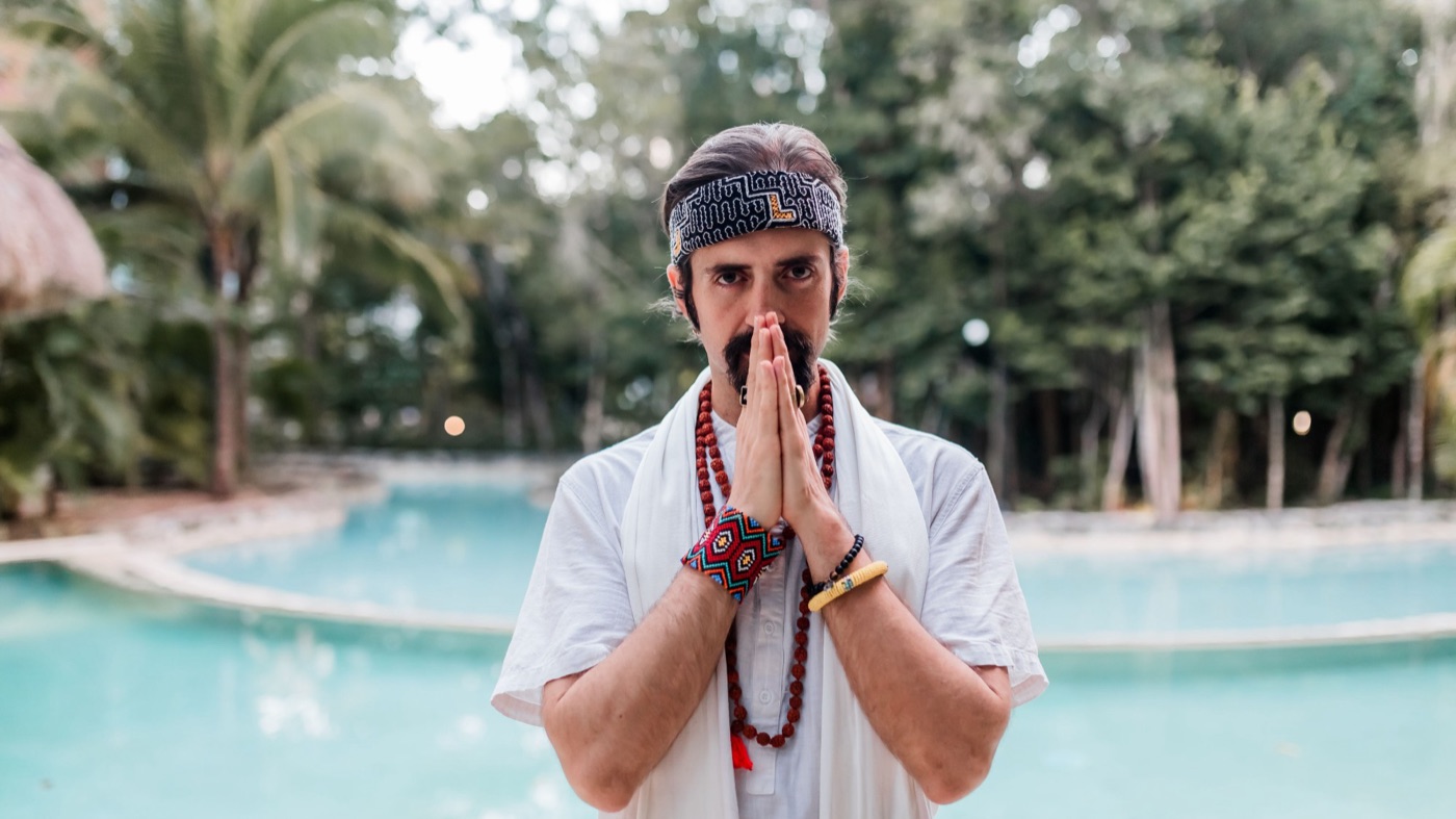 Shaman Peter in white robes and a mala, hands in prayer, before a turquoise pool in the jungle