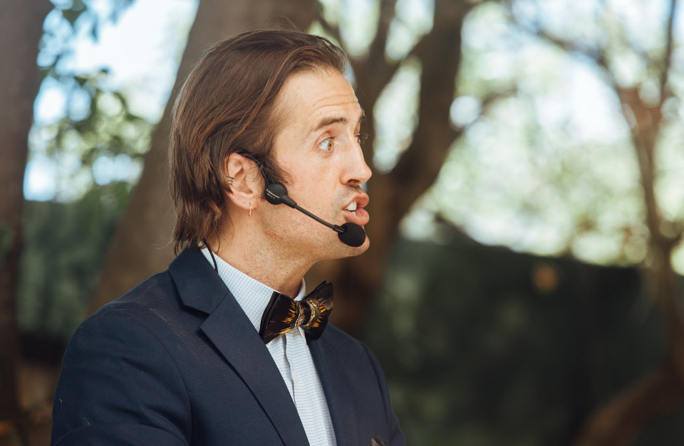 Shaman Peter in profile, speaking outdoors beneath the trees in a navy suit and feathered bow tie